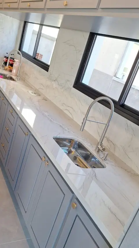 Bancada de cozinha em pedra clara com cuba de inox embutida, ao longo das janelas